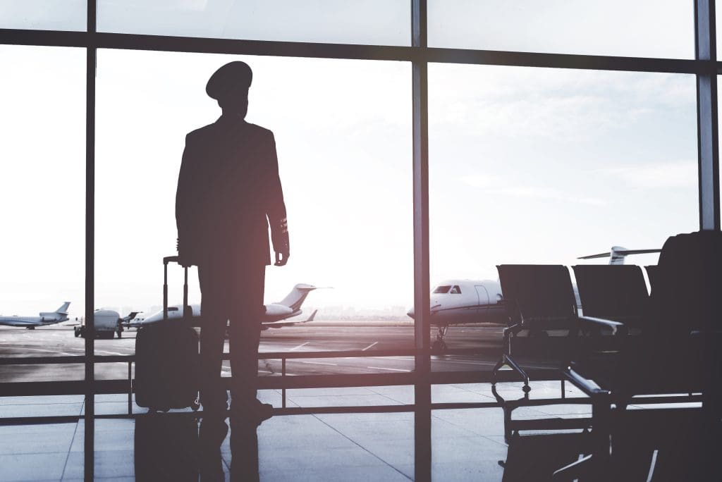 Silhouette of a pilot with a suitcase looking out an airport terminal window at private jets on the tarmac